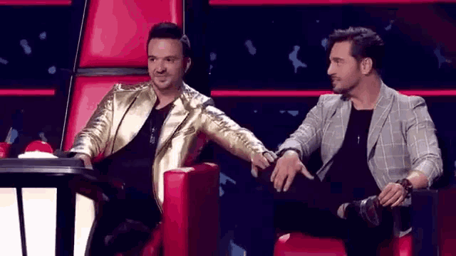 two men are sitting next to each other on a red chair and talking to each other .
