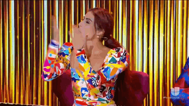 a woman in a colorful dress is blowing a kiss while sitting in front of a gold curtain .