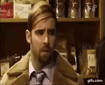a man with a beard and tie is standing in front of a shelf in a store and talking to another man .