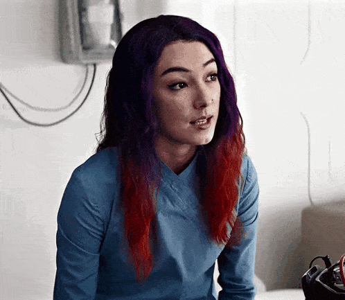 a woman with purple and red hair is wearing a blue scrub top and a blue shirt .