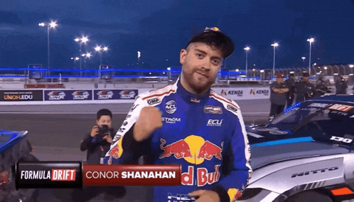 a man wearing a red bull shirt stands in front of a formula drift car