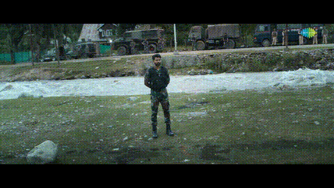 a man in military uniform stands in front of a river with military vehicles in the background