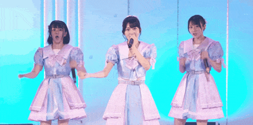 three girls are singing into microphones on a stage and one of them is wearing a blue and white dress