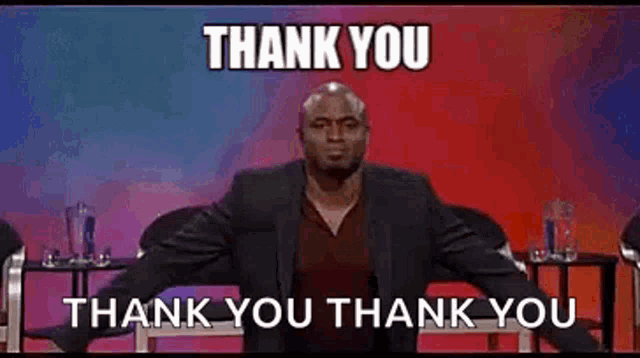 a man in a suit is sitting on a stage with his arms outstretched and saying `` thank you '' .