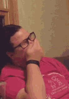 a man wearing glasses and a red shirt is sitting on a couch with his hand in his ear .