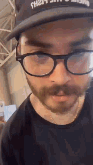 a man with a beard and glasses is wearing a black hat .