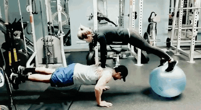a man and a woman are doing push ups in a gym while a woman stands on an exercise ball .