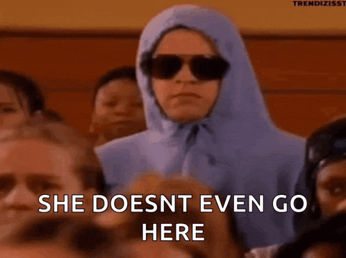 a woman wearing sunglasses and a hooded jacket is sitting in a crowd of people and says she does nt even go here