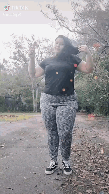 a woman in a black shirt and grey pants is standing in a park with trees in the background .