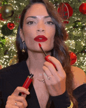 a woman applies red lipstick in front of a christmas tree