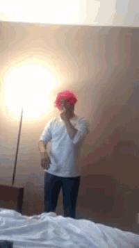 a man in a pink wig is standing in front of a lamp in a bedroom .