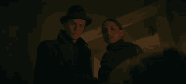 a man in a hat stands next to a woman in a dark room