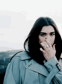 a woman wearing a trench coat and a ring is covering her nose with her hand .