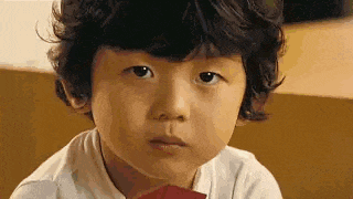 a young boy with curly hair is looking at the camera while holding a piece of paper .