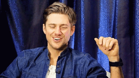a man in a denim shirt is laughing with his eyes closed and his hand up in front of a blue curtain .