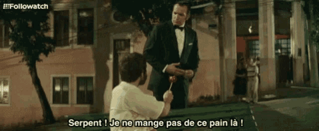 a man in a tuxedo is giving a lollipop to another man who is sitting on a park bench