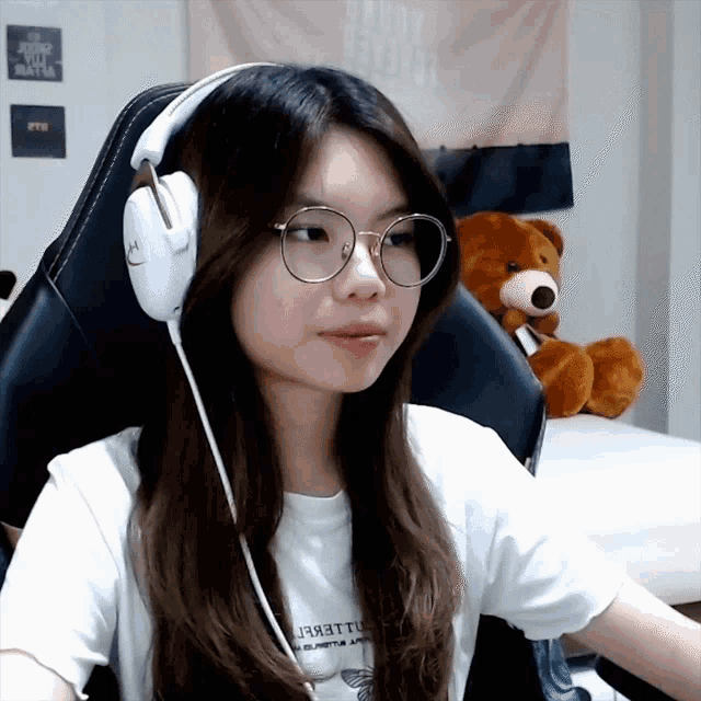 a girl wearing glasses and headphones has a teddy bear in the background