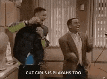 two men are dancing in a kitchen with the words `` cuz girls is playahs too '' written on the bottom .