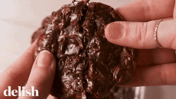 a close up of a person holding a chocolate cookie with the word delish on the bottom right