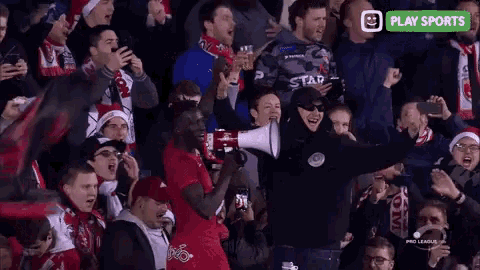 a man in a red shirt is holding a megaphone in front of a crowd of people with a play sports logo in the corner