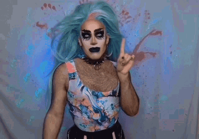 a drag queen with blue hair and black makeup is making a point with her finger .