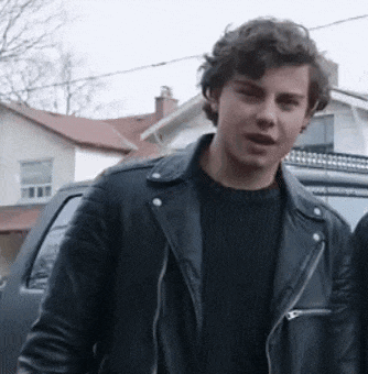 a young man wearing a black leather jacket and a black sweater is standing next to a car .