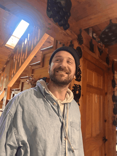 a man with a beard wearing a denim shirt and a beanie smiles