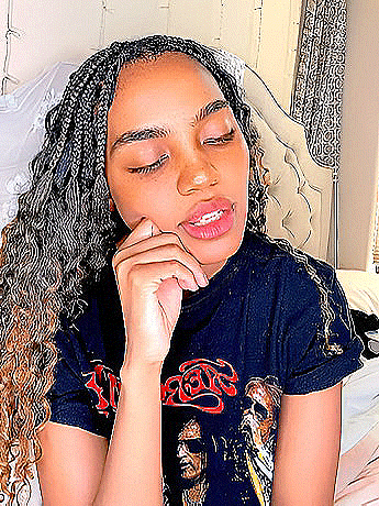 a young woman with braids is sitting on a bed with her eyes closed and her hand on her chin .