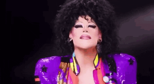 a drag queen is wearing a purple jacket and earrings and making a face .