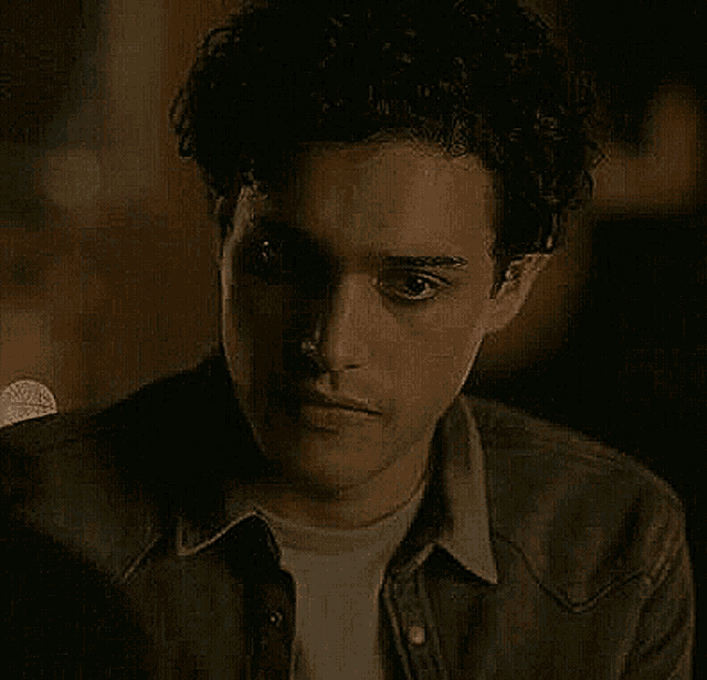 a man with curly hair is wearing a denim jacket and a white shirt and looking at the camera .