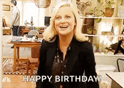 a woman is standing in a living room and smiling while saying `` happy birthday ! ''