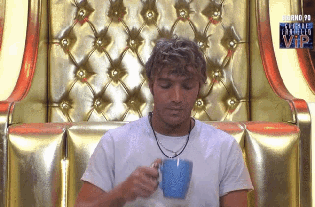 a man drinking from a blue mug in front of a gold chair with giorno 90 written on it