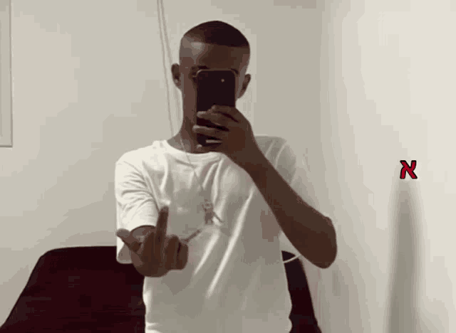 a man in a white shirt is taking a picture of himself with his phone and giving the middle finger