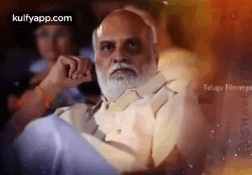 a man with a beard is sitting in a chair with his hand on his chin and looking at the camera .