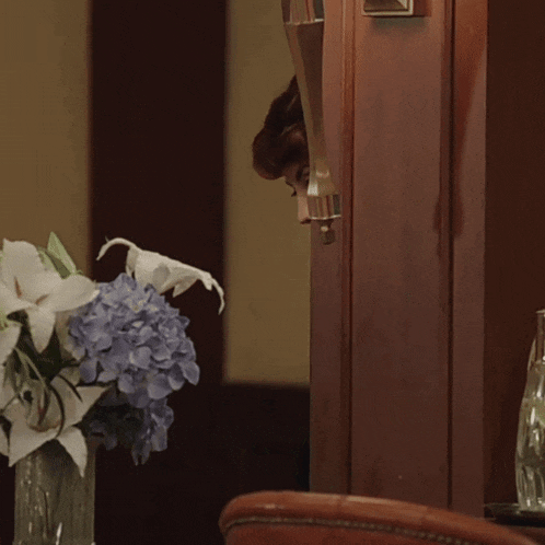a woman peeking out from behind a door next to a vase of blue and white flowers