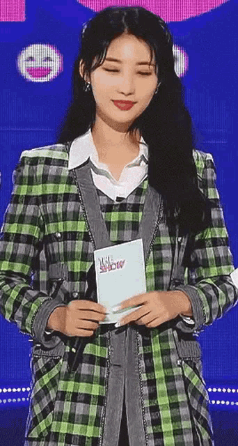 a woman in a green and gray plaid jacket is holding a microphone and a clipboard .