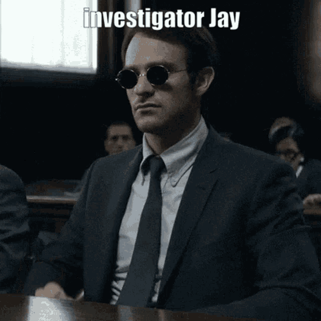 a man in a suit and tie is sitting at a table in a courtroom wearing sunglasses .