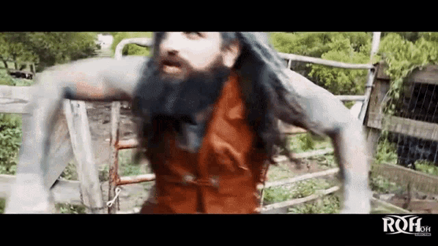 a man with long hair and a beard is standing in front of a fence with the word roh written on the bottom
