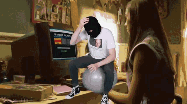 a man in a ski mask sits on an exercise ball in front of a computer screen which says feature films