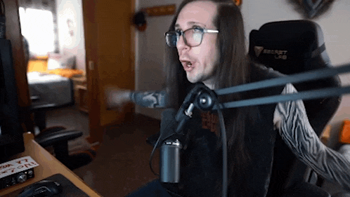 a man with long hair and glasses is sitting in front of a microphone with a secret lab chair in the background .