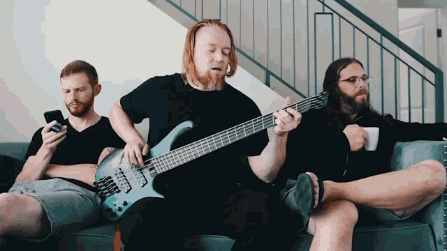 three men are sitting on a couch one of them is playing a bass guitar
