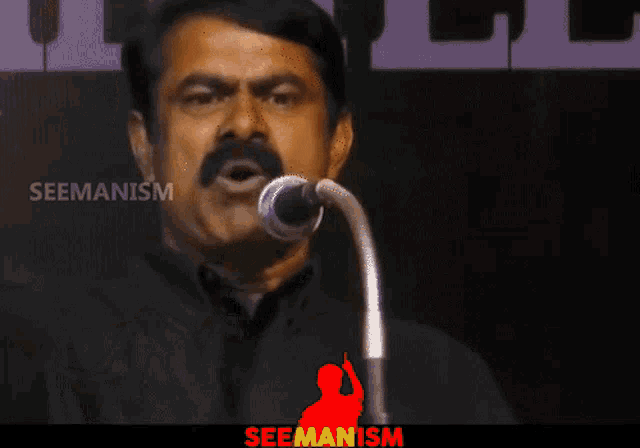 a man with a mustache is speaking into a microphone with the words seemanism on the bottom right