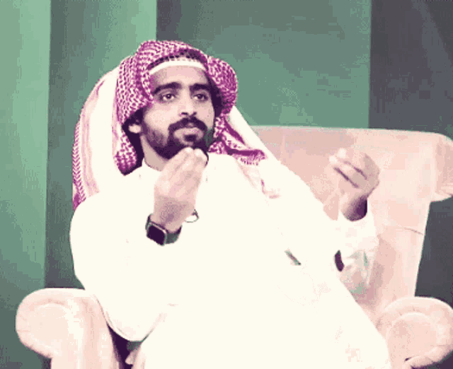 a man wearing a head scarf sits in a chair with his hands folded