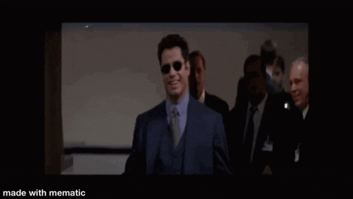 a man in a suit and sunglasses is standing in front of a group of men in suits and ties .
