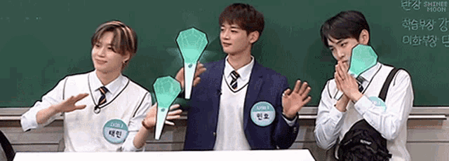 three young men in school uniforms are standing in front of a chalkboard holding glow in the dark sticks .