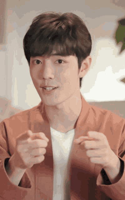 a young man wearing a brown jacket and a white shirt making a heart shape with his hands