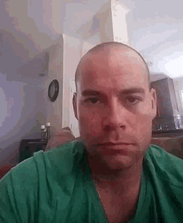 a bald man in a green shirt is sitting on a couch looking at the camera .