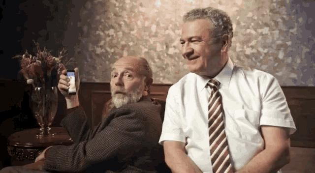 two men are sitting on a couch one of them is holding a cell phone