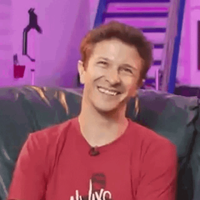 a man is smiling while sitting on a couch wearing a red shirt .