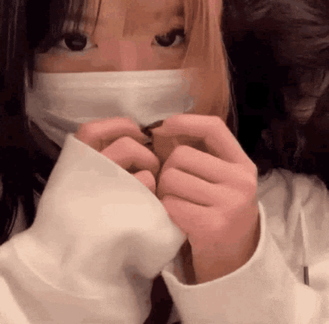 a girl wearing a mask and a white sweater is making a heart with her hands .
